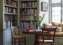 A cozy room with a wooden table and two chairs placed in front of a large window. The table is adorned with a vase of fresh flowers and books.