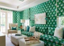 Bold blue and green print wallpaper complement a green and blue sofa accented with ivory and green pillows and matched with two ivory ottomans and a cream wingback chair. An abstract art piece hangs above the sofa between green sconces.