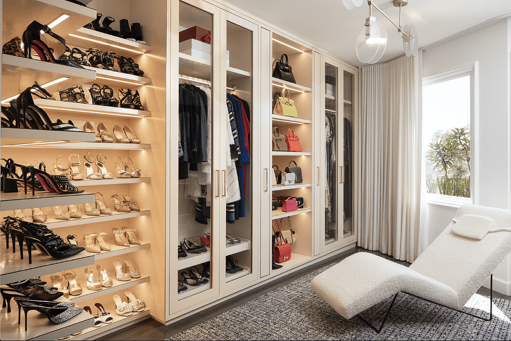 Pink-beige, l-shaped closet shoe shelves are mounted in a corner beside glass front wardrobe cabinets flanking light bag shelves. A white boucle lounge chair sits on a gray rug facing the wardrobes.