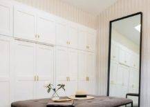 Chic walk-in closet features stacked white wardrobe cabinets, walk-in pantry skylight, beige stripe wallpaper, black metal frame leaning mirror and a brown leather tufted ottoman.