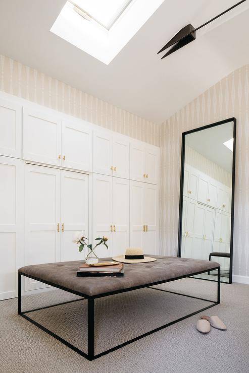 Chic walk-in closet features stacked white wardrobe cabinets, walk-in pantry skylight, beige stripe wallpaper, black metal frame leaning mirror and a brown leather tufted ottoman.