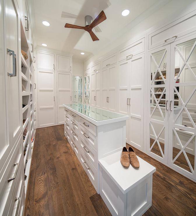 Long and narrow custom walk in closet features white cabinetry with a white built in island showcasing nickel hardware and a glass countertop. Mirrored closet doors bring an elegant touch to the interior design finished with X trim.