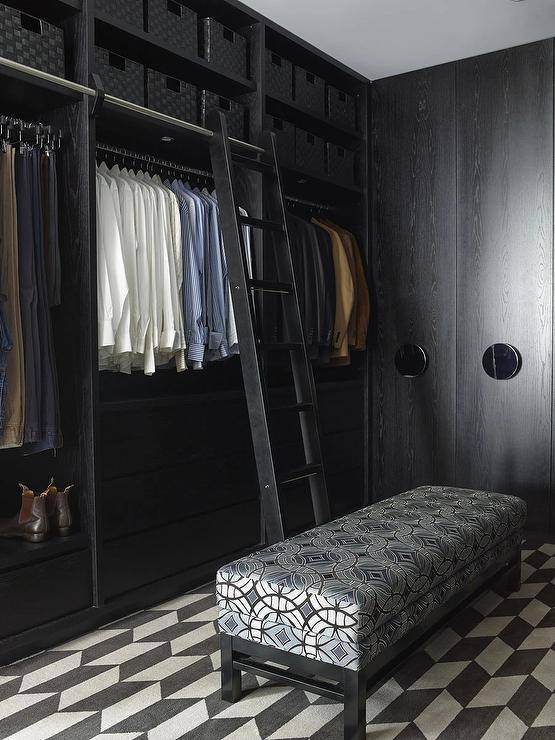 Modern black oak custom walk in closet with a black ladder on rails features cubbies with black woven baskets and a black and silver bench on black and white flooring.