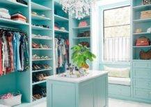 Turquoise blue walk-in closet features a window seat bench under a window surrounded by blue built-ins with glass knobs. This large walk-in closet features shoe shelves, purse shelves, and a marble top island illuminated with a stunning French crystal chandelier.