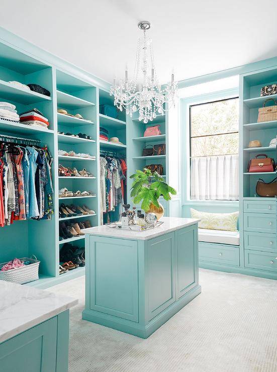 Turquoise blue walk-in closet features a window seat bench under a window surrounded by blue built-ins with glass knobs. This large <yoastmark class=