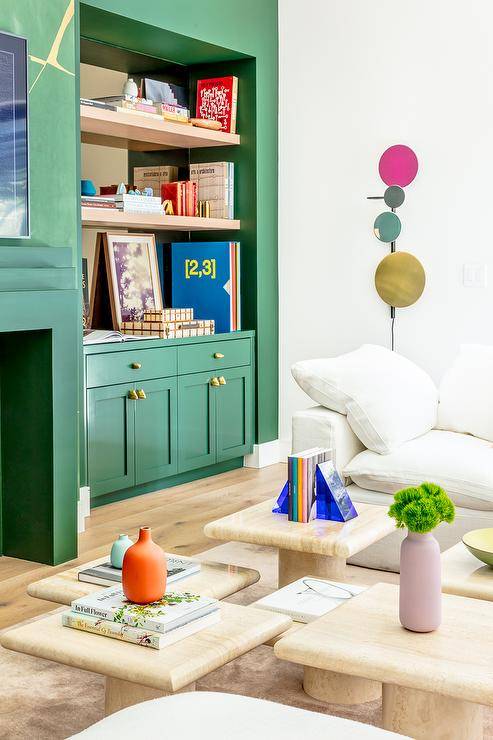 Green shaker lacquered living room cabinets adorned with brass knobs and fitted beneath inset blond wood floating shelves mounted against a mirrored backsplash and beside a green art deco fireplace. Staggered blond wood tables sit on a rug in front of a white sectional.