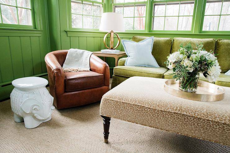 Gorgeous green and brown living room boasts windows accented with green moldings and framed by green tongue and groove walls. A white elephant sits beside a brown leather club chair lit by a Metal Banded Table Lamp placed on an end table positioned next to a green velvet skirted sofa topped with powder blue pillow. The sofa faces a gold upholstered animal print bench complemented with a round gold tray.