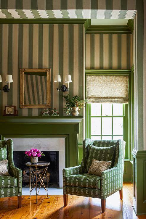 This gorgeous green formal living space features A green fireplace mantel framing a marble surround and fixed against a wall clad in green vertical stripe wallpaper. A gold French mirror hangs above the fireplace and between oil rubbed bronze 2-light sconces. A brass directoire accent table is flanked by green wingback chairs.