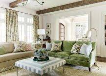 Beneath a ceiling accented with rustic wood beams, a green velvet sofa topped with blue and green pillows placed on a beige and green rug facing a blue concentric ottoman. Botanical print curtains cover French doors and windows located behind a tan sofa.