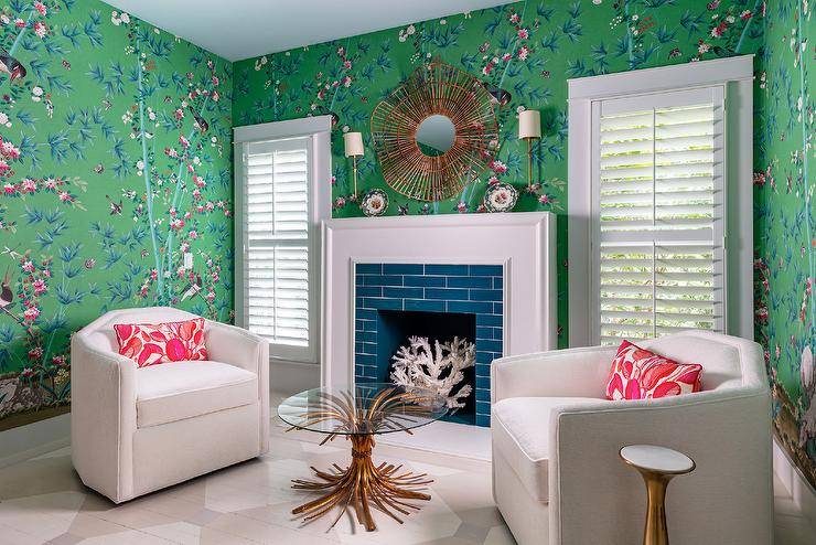 Lit by Dauphine Sconces, a wicker mirror hangs from a wall covered in green chinoiserie wallpaper over a white fireplace mantel framing blue surround tiles. The fireplace is flanked by windows covered in white shutters. White art deco swivel chairs sit on a geometric painted floor on either side of a glass and brass coffee table.