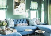 Stylish green and blue family room boasts stunning glossy green walls holding an art piece between windows covered in green roman shades and over a blue u-shaped sectional topped with blue pillows. The sectional sits on a blue ikat rug facing marble and brass hexagon accent tables.