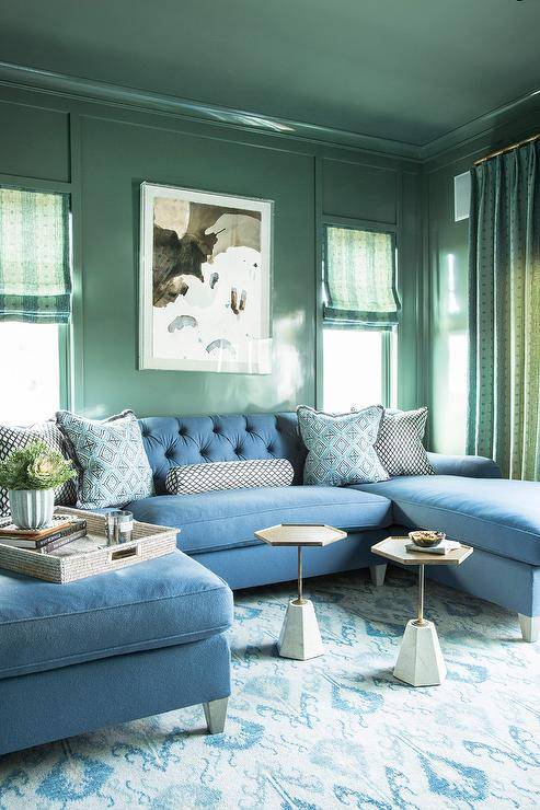 Stylish green and blue family room boasts stunning glossy green walls holding an art piece between windows covered in green roman shades and over a blue u-shaped sectional topped with blue pillows. The sectional sits on a blue ikat rug facing marble and brass hexagon accent tables.