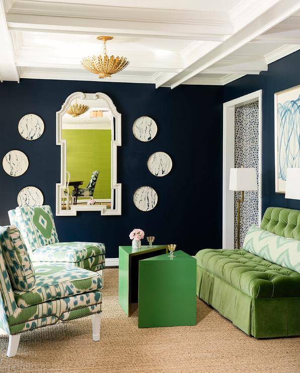 A gold chandelier hangs from a coffered ceiling over two greek ikat chairs placed on a sisal rug angled towards green triangle accent tables paired with a green velvet tufted settee. The settee is topped with a greek ikat lumbar pillows and located beneath a blue and green abstract art piece hung from a black painted wall. An adjacent wall holds a white mirror between decorative marble plates.