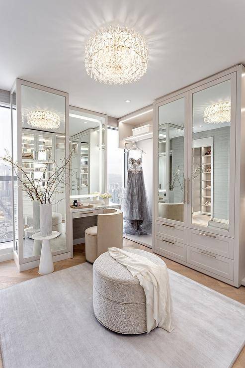 Glam light gray walk in closet illuminated by a crystal flush mount light features mirrored wardrobe cabinets, a round gray ottoman and a custom makeup vanity with cream velvet vanity chair.
