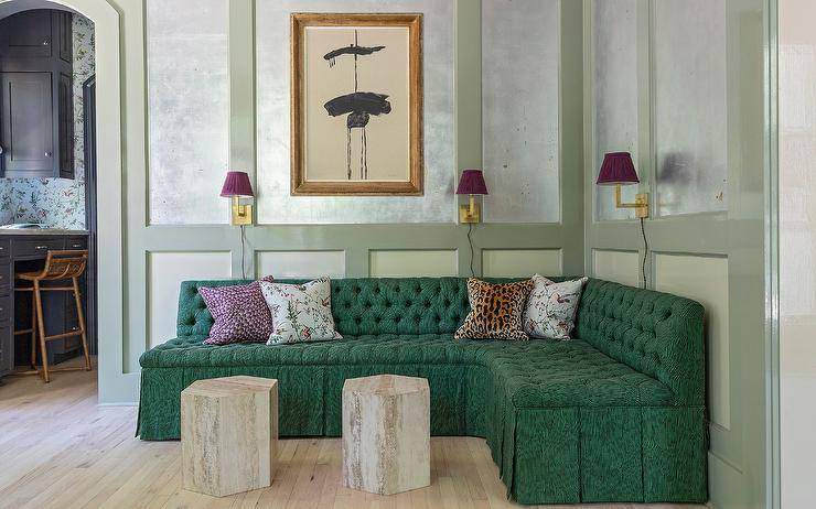 Eclectic living room features an emerald green tufted French sectional on sage green wall trim lit by scones with purple shades and dual hexagon accent tables.