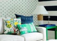 Green and blue pillows sit atop a white couch placed in front of a wall covered in green plus print wallpaper, as a wicker lamp and a blue tray sits on a green parsons end table placed beside a white and black striped accent wall.