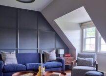 Attic family room features a blue couch on a blue paneled accent wall and round blue and brown tray coffee tables atop a blue rug and a blue accent chair.
