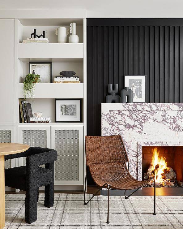 A marble slab fireplace mantel with a marble slab surround is fixed against a black vertical plank accent wall. Chunky white styled shelves are fixed over built-in cabinets.