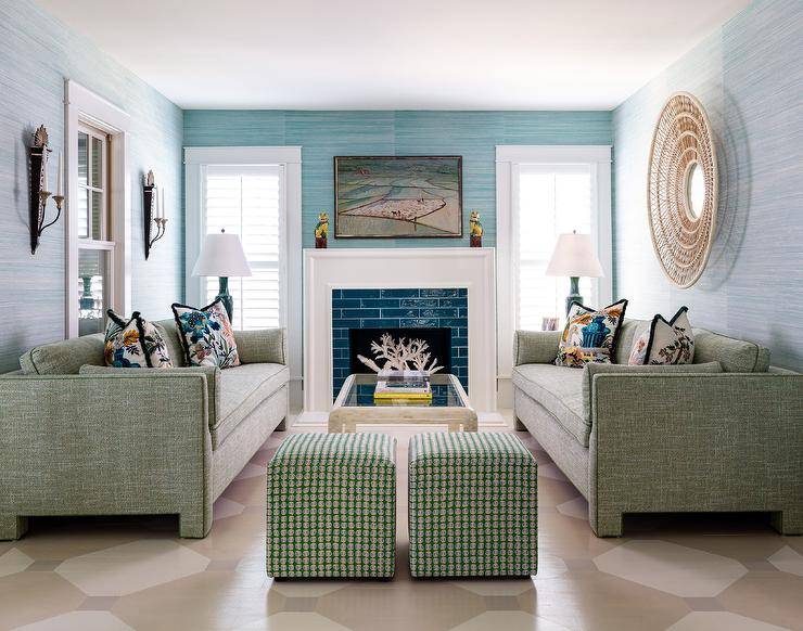 Glossy blue tiles are framed by a white fireplace mantel fixed beneath an art piece hung from an accent wall covered in blue grasscloth wallpaper. The fireplace is flanked by windows covered in white plantation shutters. A large round rattan mirror hangs from a wall clad in light blue grasscloth wallpaper over a green sofa placed on a tan geometric painted floor. The sofa faces a matching green sofa and is complemented with green cube stools.