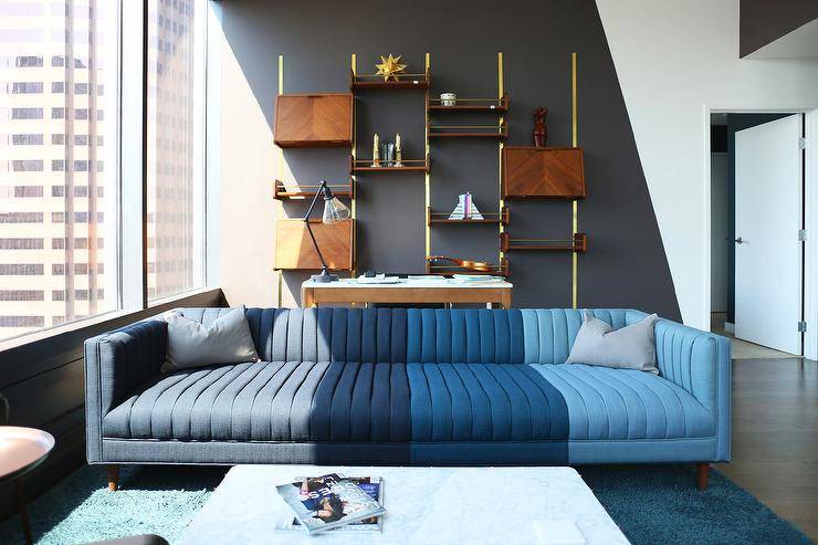 High rise apartment boasts a wonderfully designed contemporary living room featuring a blue channel tufted sofa placed on a blue rug in front of a desk positioned against brass and wood ladder shelves finishing a white and black painted accent wall.