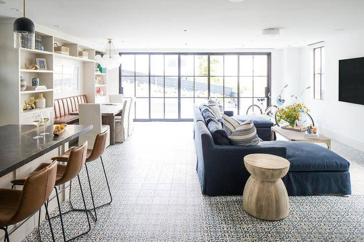 Gorgeous open plan home boasts a living room furnished with a dark blue sectional fitted with two chaise lounges and topped with white and blue striped pillows. The sofa sits on ivory and blue mosaic floor tiles beside a wooden accent table and facing a gray wood coffee table.