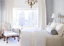 In this romantic white and gray bedroom, ivory curtain cover windows allowing natural light to hit a metal 4 poster bed dressed in white bedding accented with an ivory trim and complemented with a gray bolster pillow. The bed sits on a gray rug in front of a blue-gray grasscloth wallpapered wall lit by an eye-catching chandelier and black and white marble urn lamps.