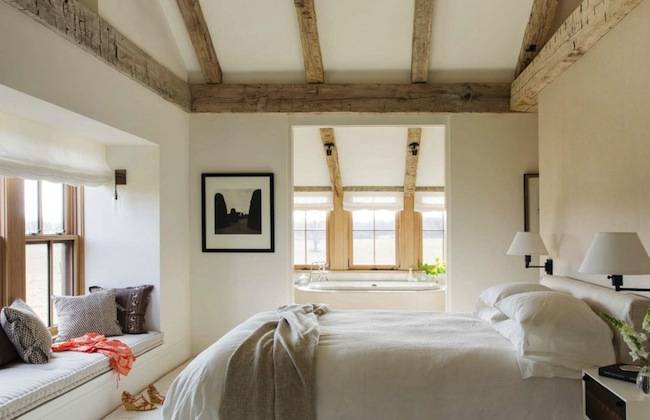 Cottage bedroom boasts a vaulted ceiling accented with rustic wood beams over a taupe headboard on bed dressed in white and taupe bedding flanked by two tone nightstands facing a window seat nook filled with pillows.