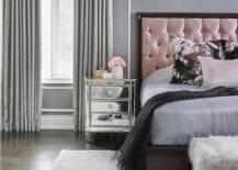 Romantic pink and gray bedroom designed with a pink and wood trim tufted headboard against a gray wall with white trim and dentil crown molding. This contemporary bedroom features a mirrored nightstand, a faux fur bench at the foot of the bed and a gray area rug that introduces sophisticated finishings to the bedroom design.