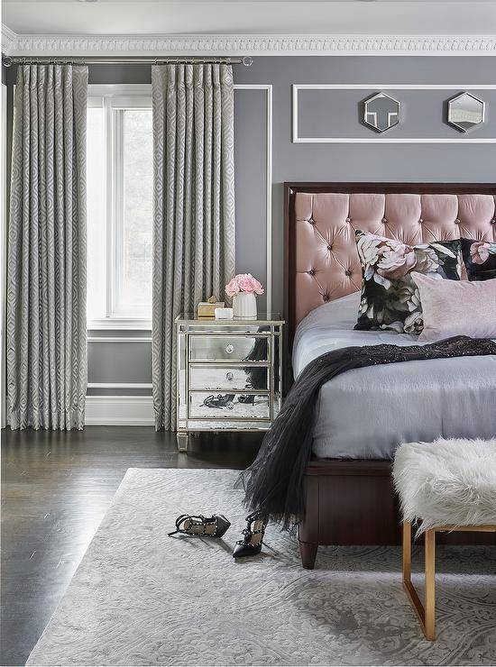 Romantic pink and gray bedroom designed with a pink and wood trim tufted headboard against a gray wall with white trim and dentil crown molding. This contemporary bedroom features a mirrored nightstand, a faux fur bench at the foot of the bed and a gray area rug that introduces sophisticated finishings to the bedroom design.