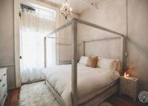 Romantic bedroom features a distressed gray canopy bed dressed in a soft white duvet illuminated by a small crystal chandelier flanked by bone inlay nightstands atop a fluffy white sheepskin rug.