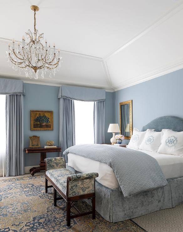 Sophisticated blue master bedroom features a blue velvet headboard that supports a bed dressed in a blue velvet pleated skirt, and white and blue monogrammed bedding, atop layered bedroom rugs. The bedroom boasts blue curtains and a gold Greek key mirror over a white gourd lamp. At the foot of the bed is a blue and white bench under a chandelier.