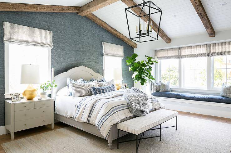 A Darlana Lantern hangs from a vaulted shiplap ceiling accented with rustic wooden beams and is positioned above a black bench finished with a white cushion. The bench sits on a gray rug in front of a light gray wooden bed covered in white and blue bedding complemented with layered white and blue pillows. The bed is positioned against a wall covered in blue grasscloth wallpaper and between white dresser nightstands topped with gold leaf lamps. The nightstands sit beneath windows fitted with gray and white striped roman shades. The room is completed with a white built-in window seat paired with a blue seat cushion.