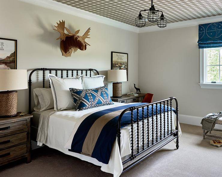 Well designed cottage bedroom boasts gray walls complementing a black and white plaid wallpapered ceiling holding an industrial cage chandelier in front of a black spindle bed. The bed is accented with white and blue hotel bedding and placed beneath a cardboard moose head and between rustic wood nightstands lit by rope spindle lamps placed in front of hung art.