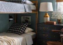 Shared boys' bedroom features a rustic bunk bed dressed in black and white bedding complemented with black plaid pillows. Black swing arm sconces light each bed, as art hangs from blue walls. A white and mustard yellow lamp sits on a black dresser finished with brass hardware and positioned beneath a window dressed in a bamboo roman shade. A wooden chair sits on a green plaid rug.