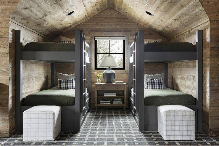 Attic boy's bunk bedroom features charcoal gray bunk beds with olive green bedding, a shared wooden nightstand lit by a gray lamp, an oak plank ceiling with oak plank walls, cube stools at the foot of beds and a charcoal gray rug.