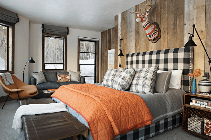 Welcoming country bedroom boasts a gorgeous black and white gingham bed complemented with an orange quilt and black plaid pillows. The bed sits on a gray rug against a wall covered in rustic wood vertical panels. A decorative animal bust is mounted above the headboard, while the bed is lit by oil rubbed bronze swing arm sconces fixed over wooden nightstand with metal grill accents.