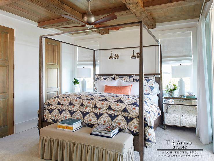 Welcoming cottage bedroom features a ceiling fan hung from a rustic wood coffered ceiling over a brown wood canopy bed dressed in taupe and blue bedding topped with ab orange bolster pillow. The bed is illuminated by two swing arm sconces mounted between windows located above antique mirrored nightstands lit by white ceramic lamps. In front of the bed, a beige skirted bench sits on light gray carpeting.
