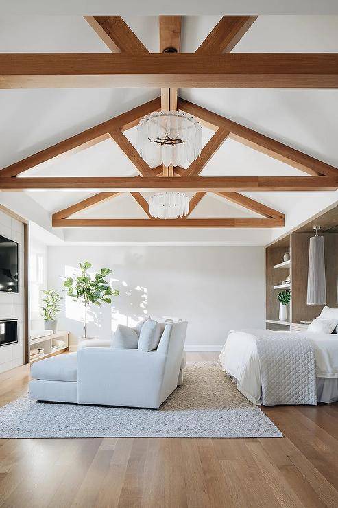 A glass icicles chandelier hangs from a truss ceiling in an elegant master bedroom boasting white his and hers loungers placed on a light gray rug facing a fireplace finished with a TV.