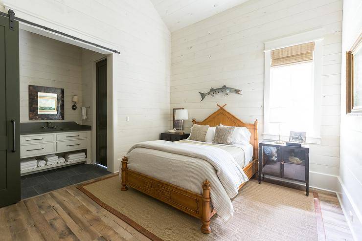Charming rustic cottage bedroom features a fish wall mount fixed to a white shiplap wall over a faux bamboo bed placed on a brown bound sisal rug and between mismatched industrial nightstands. One nightstand is positioned beneath window covered in a bamboo roman shade. A black barn door on rails slides open to reveal an ensuite bathroom.