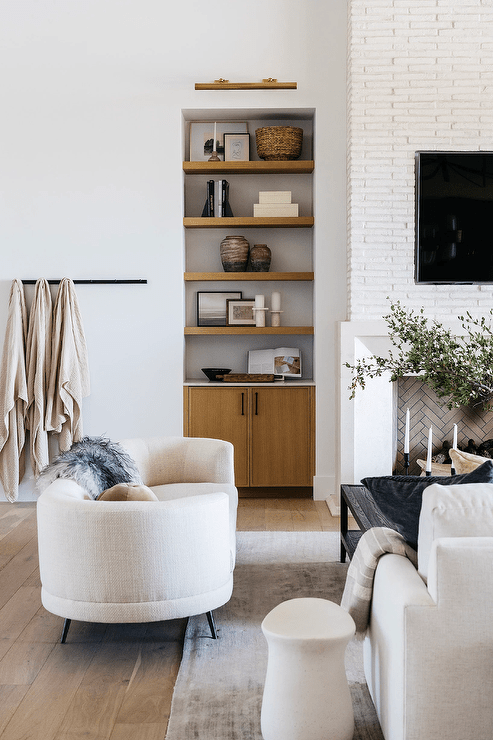 Living room features a white curved sofa accented with a beige velvet tufted ball pillow, a wall mount throw blanket rack and a built in shelf next to a fireplace.
