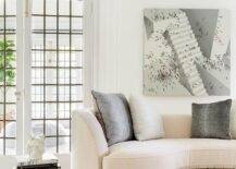 Art hangs from a white wall accented with white moldings over a cream velvet curved sofa topped with gray pillows. Stacked coffee table books sit on a gray rug beside the sofa.