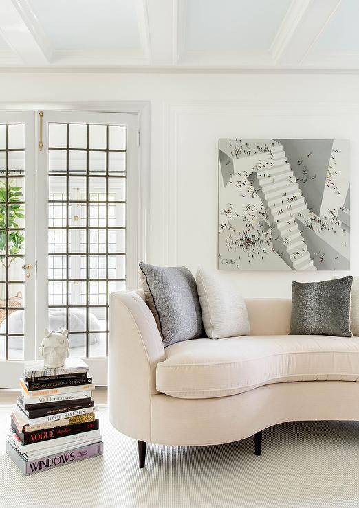 Art hangs from a white wall accented with white moldings over a cream velvet curved sofa topped with gray pillows. Stacked coffee table books sit on a gray rug beside the sofa.