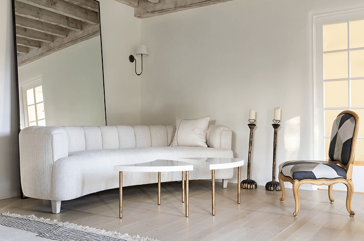 Living room features a white curved channel tufted sofa in front of a full length mirror, white and gold accent tables, a white, black and gray French chair and tall candle floor stands.