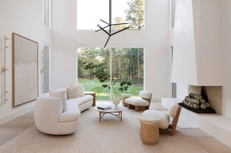Modern living room features a white boucle curved sofa with round marble top coffee table, a modern fireplace design and low wood and fabric accent chairs atop a light brown jute rug.