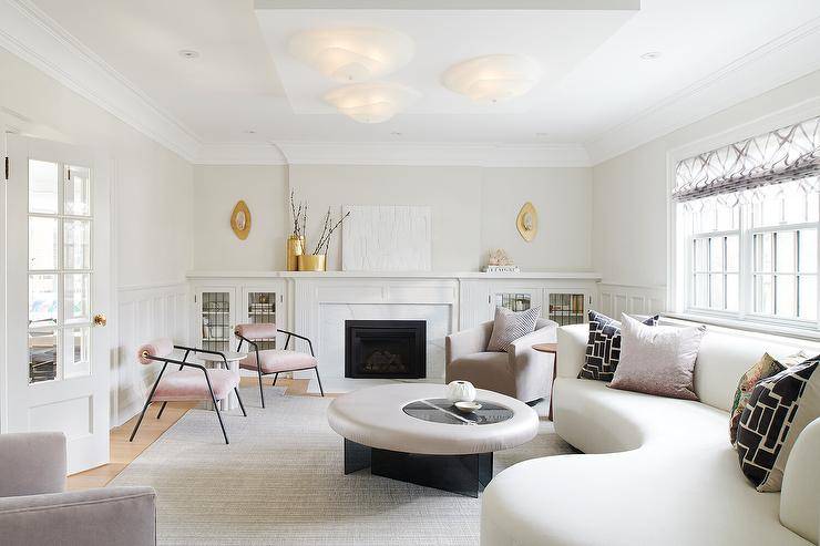 Luxurious living room features a white curved sofa placed beneath windows and topped with black and silver pillows. The sofa sits on a light gray rug at a round coffee table.