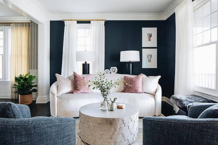 Gorgeous curved white sofa boasts pink velvet greek key pillows, a with round silver honeycomb coffee table, atop a white and gray rug. Two blue denim barrel back accent chairs and windows covered in white curtains complete this stunning living room.