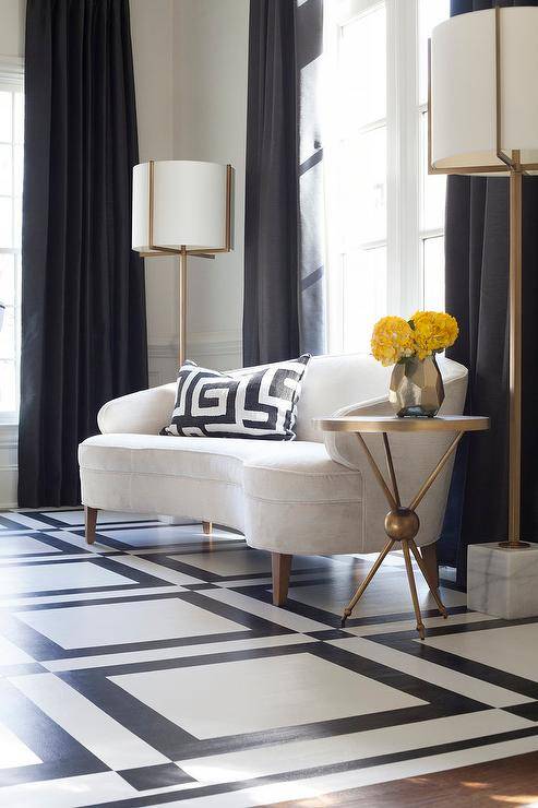 Clad in black and white geometric floor tiles, this stunning dining room features a Mitchell Gold + Bob Williams Vera Sofa topped with a black and white pillow and positioned beside a marble and brass accent table in front of a window dressed in black curtains lit by Arteriors Pearson Floor Lamps.