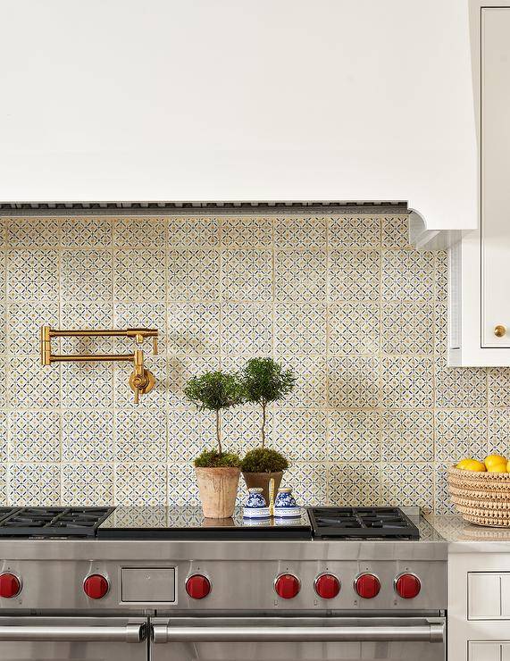 A stainless steel range sits against ivory, orange, and black mosaic backsplash tiles fitted with an aged brass swing arm faucet mounted under a white hood.