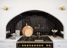 Kitchen features an arched cooking nook with glossy black grid tiles, a brass swing arm pot filler over a gold and black stove and white cabinet drawers with brass knobs and a marble countertop.