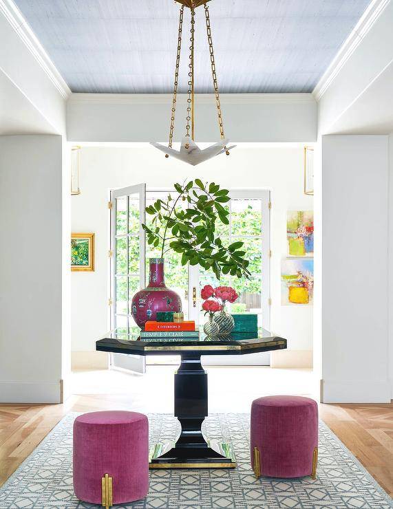 Luxurious entryway features a gold and black octagon table paired with fuchsia stools and placed on a white and blue rug beneath a blue tray ceiling fitted with a white and gold pendant.